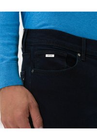 Dark blue jeans featuring a small, white BRAX label on the pocket. The fabric appears smooth with a subtle texture. The waistband is fastened with a button.