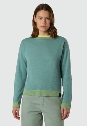 Donna che indossa un maglione a maniche lunghe di colore turchese chiaro con rifiniture giallo-verdi e pantaloni verde pallido, in piedi davanti a uno sfondo grigio semplice.