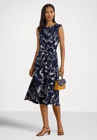 Marineblå kjole med hvitt blomsterprint, ermeløs design, vridd detalj i midjen; modellen holder en liten beige kurvveske med marineblå aksenter.