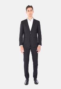 Completo nero con giacca a un bottone, rever a forma di punta e pantaloni con tasche piatte. Camicia bianca sotto, abbinata a scarpe eleganti nere.