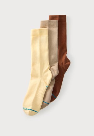 Tre ribstrikkede ankelsokker i creme, beige og brun, stablet diagonalt på en hvid baggrund.