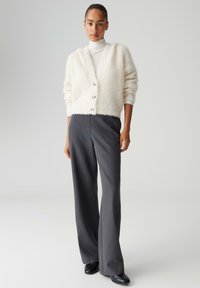 Cremefarbener, strukturierter Strickcardigan mit V-Ausschnitt und drei Knöpfen, kombiniert mit hoch taillierter, weit geschnittener grauer Hose und schwarzen Schuhen.