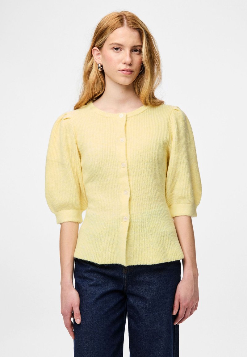 Jauna sieviete valkā gaiši dzeltenu poga priekšpusē ar pūkainām pusgarām piedurknēm un tumši zilas džinsas uz vienkārša fona.