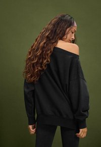 Felpa nera oversize con scollatura asimmetrica, polsini e orlo a costine, abbinata a leggings neri. Capelli castani ondulati che scendono.