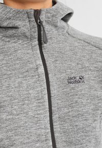 Szara polarowa kurtka na zamek z wysokim kołnierzem i haftowanym logo Jack Wolfskin na klatce piersiowej, noszona przez osobę.