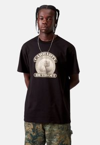 Czarna bawełniana koszulka z dużym okrągłym nadrukiem "Carhartt WIP Detroit". Model ma na sobie spodnie w kamuflażu, minimalną biżuterię i elegancką fryzurę.