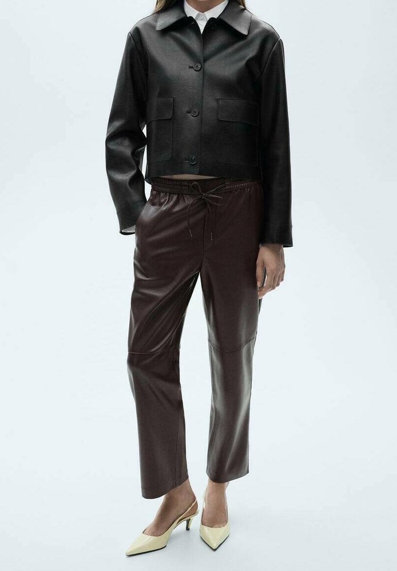 Femme portant une veste courte en cuir noir, un pantalon en cuir marron foncé avec cordon de serrage, et des escarpins crème à bout pointu sur fond blanc.