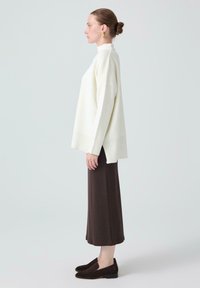 Hvid strikket sweater med høj hals, oversized pasform og ribbet kant; parret med en lang brun nederdel og mørke slip-on sko.