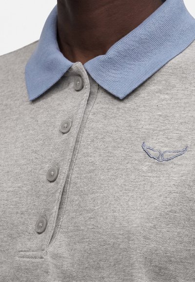 Κοντινό πλάνο ενός ατόμου που φοράει ένα ανοιχτογκρι πόλο με γαλάζιο γιακά και μικρό κεντημένο λογότυπο στο στήθος.