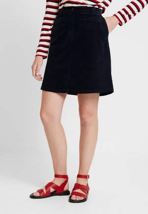 Vrouw draagt een zwarte rok tot op de knie, een rood-wit gestreept shirt met lange mouwen en rode platte sandalen met enkelbandjes.
