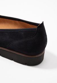 Zapato plano negro de ante con punta redondeada, un interior texturizado en crema y una suela de goma que presenta un ligero tacón y un detalle de costura visible.