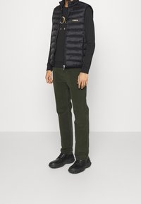 Person som bär en svart vadderad väst över en svart långärmad tröja, mörkgröna byxor och svarta snörskor, stående mot en vit bakgrund.