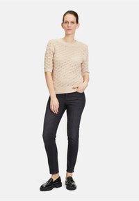 Helle beige gestrickte Sweater mit einem strukturierten Design, halben Ärmeln und gerippten Bündchen, kombiniert mit dunklen, eng anliegenden Jeans und schwarzen Schuhen.