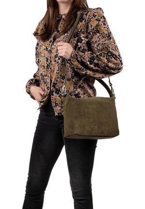Olivgrünes Wildleder-Handtäschchen in rechteckiger Form, mit verstellbarem Riemen und sichtbaren Nähten; getragen zu einer gemusterten Bluse und dunklen Jeans.