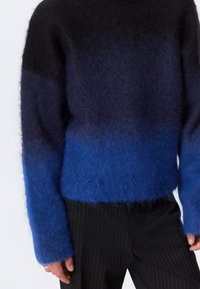 Maglione con gradiente blu e nero dalla texture morbida; presenta maniche lunghe e una vestibilità ampia. Abbinato a pantaloni a righe. 