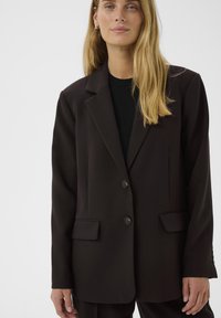Donna con capelli biondi lunghi, che indossa una giacca nera over-size sopra un top nero, in piedi contro uno sfondo chiaro e semplice.