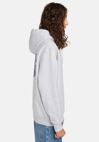 Sudadera gris con un ajuste relajado, que cuenta con un cordón, puños acanalados y un diseño bordado en la manga izquierda.