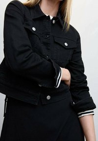 Giacca in denim nero con bottoni argentati, due tasche sul petto e polsini a righe. Presenta un design aderente e dettagli di cucitura in vista.