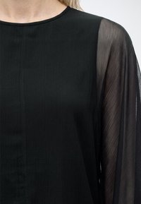 Gros plan d'un haut noir avec un devant uni et une manche longue transparente et texturée sur le côté droit.