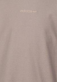 Rosa tyg T-shirt med en liten, präglad Adidas-logotyp i en ljus guldfärg, som framhäver en mjuk textur och minimalistisk design.