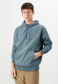 Sudadera texturizada azul claro con un bolsillo frontal, cordones ajustables y puños de canalé, llevada sobre pantalones beige.