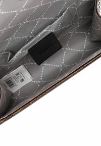 Interior de bolsa marrón con forro gris estampado. Presenta una etiqueta negra. Etiqueta de cuidado visible y un bolsillo. La textura parece suave.