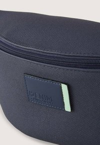Navyblauwe textuurstof tas met een gebogen bovenkant, ritsluiting en een rechthoekig leren label met reliëflettering en een mintgroene accent.