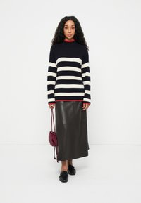 Pull en maille rayé bleu marine et blanc avec un col rouge, accompagné d'une jupe midi en cuir noir et de mocassins noirs, tenant un sac bordeaux.