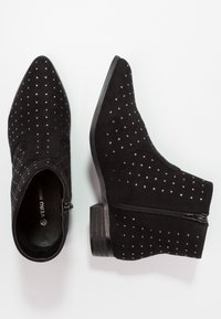Botas de tobillo de ante negro con punteras afiladas, acentos tachonados, cremallera lateral y tacones cortos en bloque. Superficie texturizada con un interior suave.