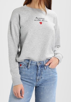 Graues Sweatshirt mit dem Schriftzug „tommy with love“ und roter Herzstickerei, kombiniert mit hellblauen Jeans, die auf der Tasche ein kleines rotes Herz haben.
