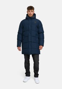 Navy blue puffer coat with a hood, featuring quilted panels, front pockets, and a logo detail. Worn over dark jeans and white sneakers.