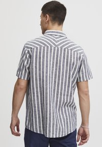 Lühikeste varrukatega särk, millel on vertikaalsed sinised ja valged triibud, nööbiga kaelus ning seljaosa chevron-mustriga detail.