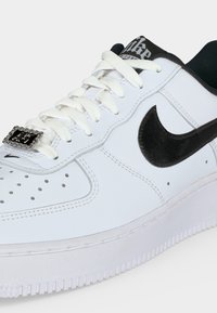 Λευκό αθλητικό παπούτσι Nike Air Force 1 με μαύρο σήμα, λευκά κορδόνια και μεταλλικό διακοσμητικό στα κορδόνια σε απλό φόντο.