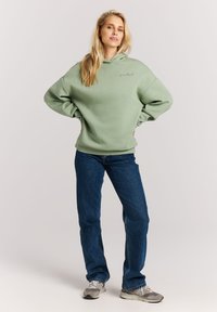 Lichtgroene hoodie met een ontspannen pasvorm, voorzien van een klein logo op de borst. Gecombineerd met blauwe jeans en grijze sneakers.