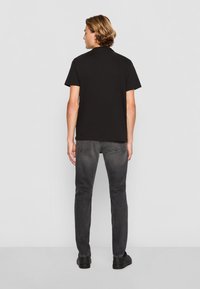 Camiseta negra de manga corta, de tela de algodón, con cuello redondo, combinada con jeans oscuros de corte ajustado y zapatillas bajas negras. Vista trasera.