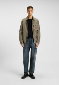 Letvægts olivengrøn jakke med en forlommeslåg og lynlås, kombineret med mørkeblå jeans og sorte sko, på en ensfarvet baggrund.