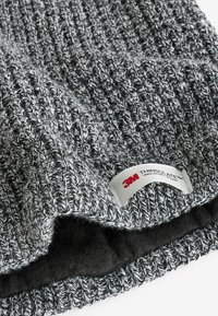 Grå strikket hue med et tekstureret mønster, der har en sort fleece-foring og et hvidt mærke med "3M THINSULATE" trykt i rødt.
