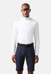 Hvid langærmet zip-trøje parret med marineblå shorts. Har lynlåslommer og et sort bælte med en metalspænde. Mørke solbriller.