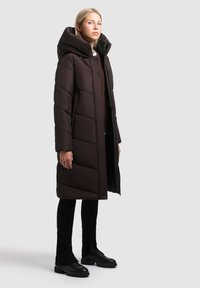 Garš melns brūns pūkains mētājs ar lielu kapuci, kam ir izšūti paneļi, priekšējā rāvējslēdzēja aizdare un sānu kabatas. Uzvilkts virs melnajām biksēm un zābakiem.