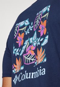 Marineblå t-skjorte med blomstergraphikk på baksiden, med blader og blomster i rosa, oransje og turkis, med "Columbia"-tekst i hvitt.