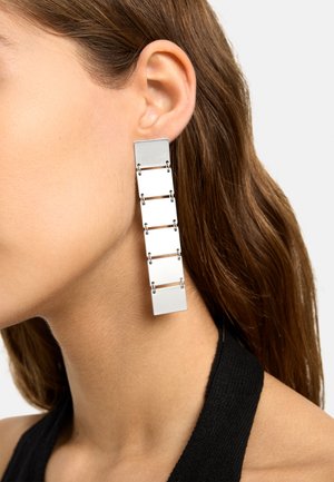 Kobieta z długimi, brązowymi włosami, nosząca na lewym uchu długą, prostokątną srebrną kolczyk wykonaną z połączonych metalowych segmentów, na białym tle.
