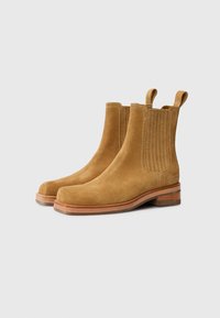 Botas de tobillo de ante color tostado con un acabado suave, paneles laterales elásticos y tiradores. Tacón plano apilado y diseño de punta redondeada.