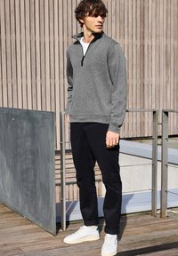 Šedá fleece mikina na zip s bílým vnitřním tričkem, kombinovaná s černými kalhotami a bílými teniskami, stojící na dřevěné terase s kovovým zábradlím.