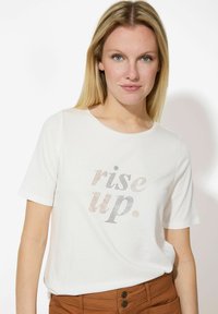 Weißes T-Shirt mit kurzen Ärmeln, auf dem der Spruch "rise up" aus Strasssteinen besteht. Rundhalsausschnitt, weicher Stoff, lässige Passform, kombiniert mit braunen Hosen.