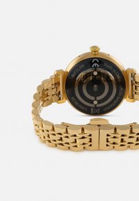 Guldtonet armbåndsur med et poleret metalarmbånd, rund urkasse og en sort urskive med cirkulære detaljer og tekst på bagsiden.