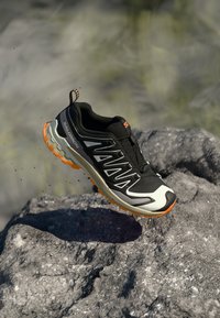 Black and white Salomon trail running shoe with orange sole hovering above rocky surface, dirt particles in the air.