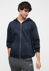 Σκούρα μπλε φούτερ με φερμουάρ, μπροστινή τσέπη, κορδόνια στο καπό και ριμπ μανσέτες, φορεμένο με γκρι παντελόνια φόρμας. Απλός, casual σχεδιασμός.