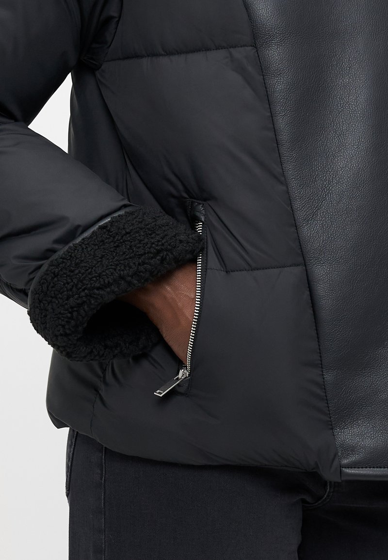 Giacca a vento nera con pannello in pelle. Presenta un polsino in pile testurizzato e una tasca con zip sul lato sinistro.