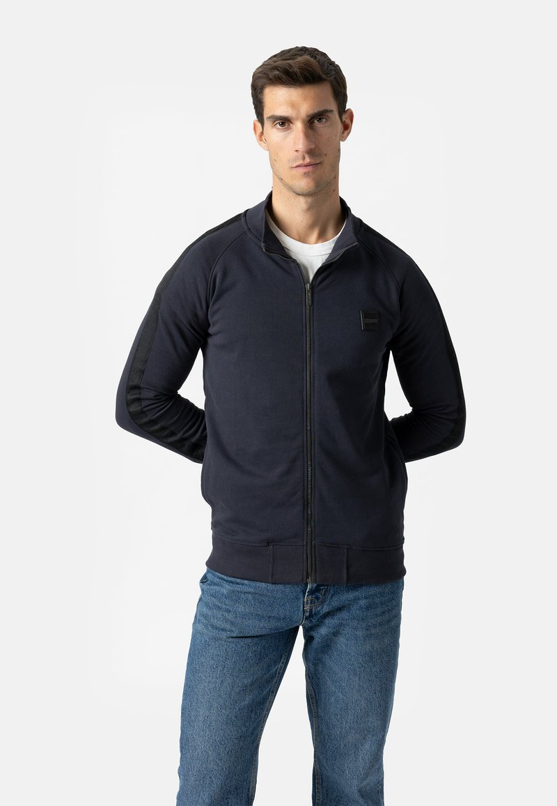 Marineblauw sweatvest met ritssluiting, een bomberkraag, geribde manchetten en accenten op het zijpaneel. Gecombineerd met lichtblauwe spijkerbroek.