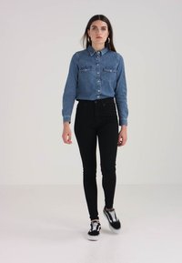 Denim srajca z gumbi v modri barvi, z dvema žepoma na prsih in ovratnikom, v kombinaciji z črnimi skinny kavbojkami in črnimi supergami z belimi detajli.